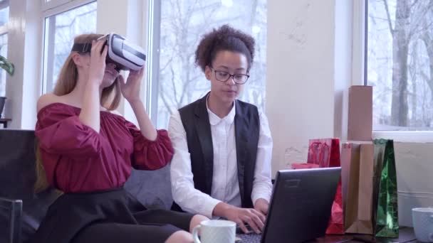 Modern teknolojiler, bilgisayarlı dizüstü bilgisayarı olan sevimli siyah kız ve Kafkasyalı kız arkadaş sanal gerçeklikte VR gözlük oyununda oturuyor. — Stok video