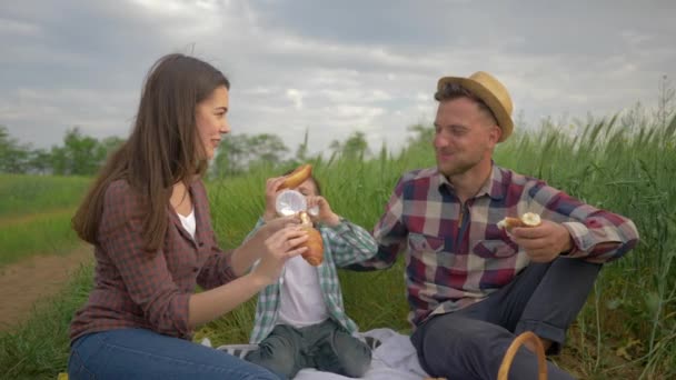 Hermosa mujer feliz alimenta al hombre mientras se relaja con el niño en el picnic familiar al aire libre en el campo verde de cerca — Vídeos de Stock