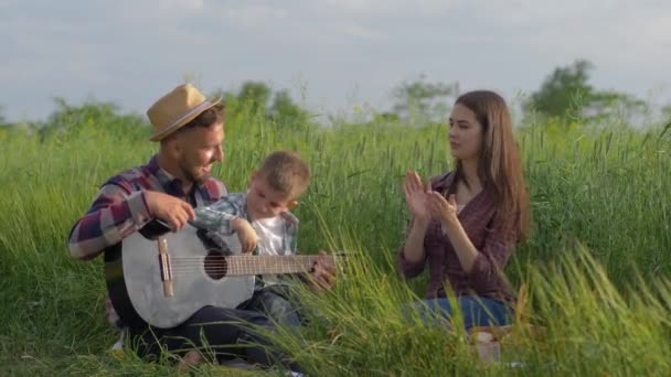 Anak laki-laki karismatik adalah belajar untuk bermain gitar sementara ibu dan ayah tertawa dan mengagumi sambil bersantai pada piknik keluarga di rumput hijau — Stok Video