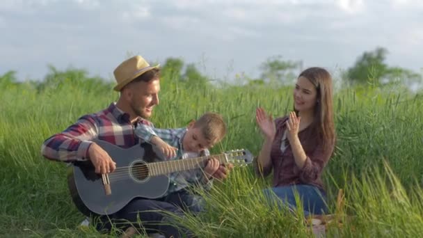 Familia creativa, feliz papá risueño enseña a su hijo a tocar el instrumento de cuerda mientras mamá aplaude y ríe mientras se relaja en el picnic en la naturaleza en el campo verde — Vídeos de Stock