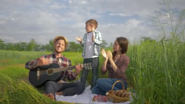 Divertida familia, alegre macho toca la guitarra mientras que la hembra con el niño baila y aplaude mientras se relaja en el picnic familiar en la naturaleza en el campo verde — Vídeo de stock