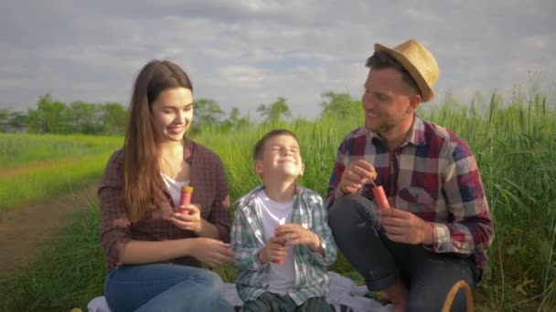 Feliz infancia, divertido padre con mamá y niño soplando burbujas y sonriendo durante las vacaciones al aire libre en el campo verde contra el cielo — Vídeo de stock