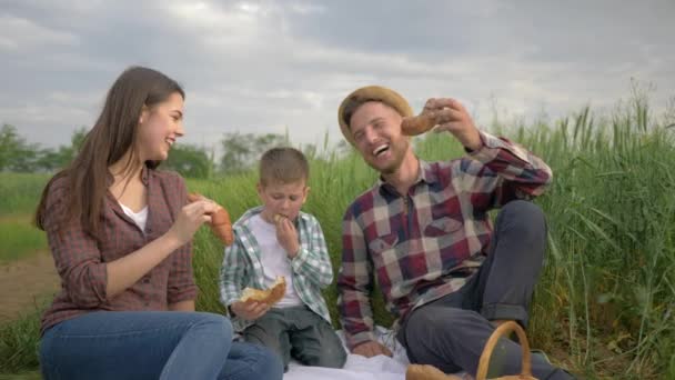 Счастливые семейные выходные, веселый мужчина кормит женщину булочкой во время отдыха на пикнике с ребенком пить молоко в зеленом поле — стоковое видео