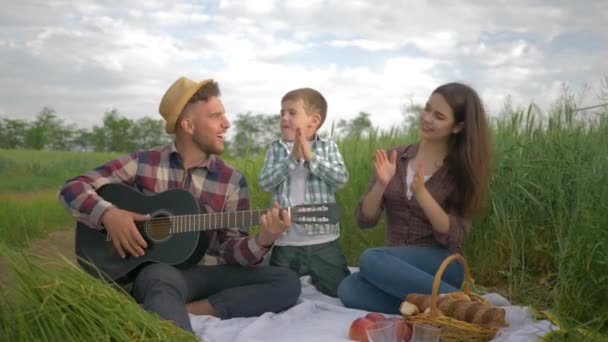 Família feliz, pai alegre toca guitarra, enquanto a mãe com o filho cantar e bater palmas enquanto relaxa no piquenique na natureza no campo verde — Vídeo de Stock