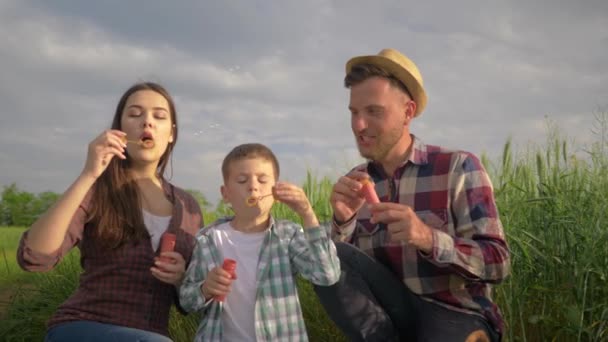 Lycklig familj, liten pojke med mamma och pappa blåser såpbubblor under roliga fritid på naturen i fält mot himlen — Stockvideo