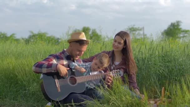 Lachen vrolijk man leert kind jongen naar gitaar spelen terwijl mam is genieten en glimlachen terwijl ontspannen op familie picknick in de natuur in groen veld — Stockvideo