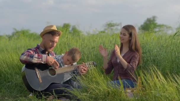Muzikale opvoeding, vrolijke grappige papa leert zoon gitaar spelen terwijl klapt en lacht terwijl ontspannen op familie picknick in de natuur in groen gras — Stockvideo