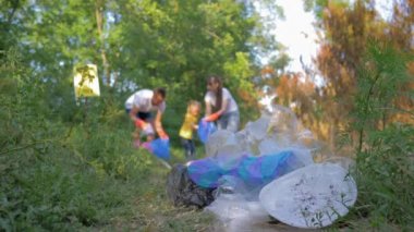 Çevresel felaket, küçük çocuklu genç gönüllüler çöp torbasında çöp toplarken, yeşil çimenlerdeki plastik ve polietilen maddeleri temizliyor.