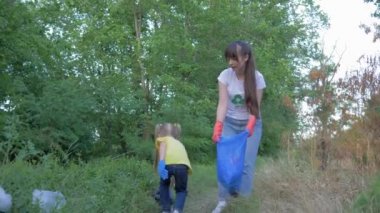 Kirliliği durdurun, küçük kız lastik eldivenli anneye çöp torbasında plastik ve polietilen çöpleri toplaması için yardım ederken yeşil çimenlerdeki işaretin yanında doğayı temizliyor.