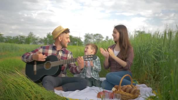 Tehetséges vidám férfi játszik vonós hangszer, míg a nő a gyerek énekelni és tapsolni, miközben pihentető piknik szabadban zöld mezőn — Stock videók