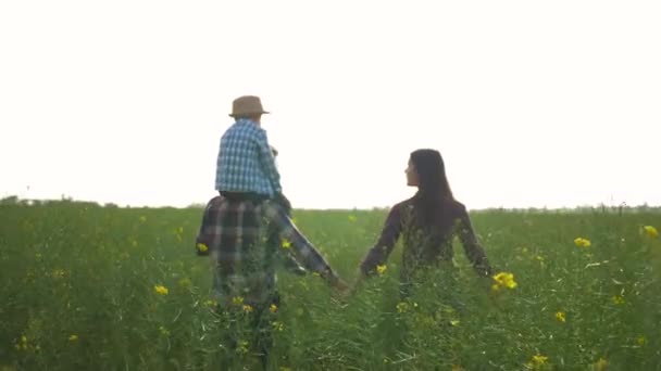Фермерство, щасливий чоловік з дитиною хлопчик на плечах і жінка ходить зі зброєю на бік в зеленому полі ріпаку проти неба — стокове відео