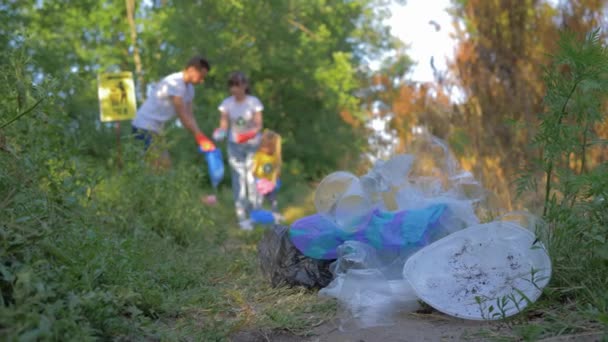 Inquinamento di plastica, ragazza e ragazzo con bambino raccoglie spazzatura in sacco della spazzatura in sfocato durante la pulizia all'aperto da plastica e polietilene in erba verde — Video Stock