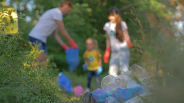 Problém domácího odpadu, matka a otec s malou dcerou sbírají odpadky do pytle na odpadky v rozostřeném stavu při čištění přírody z plastu a polyethylenu v zelené trávě — Stock video