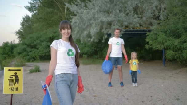 Sprzątanie plaży, portret młodej wolontariuszki matki w gumowych rękawiczkach z workiem na śmieci przy znaku wskaźnika na tle ojca i córeczki po zbieraniu plastikowych odpadków — Wideo stockowe