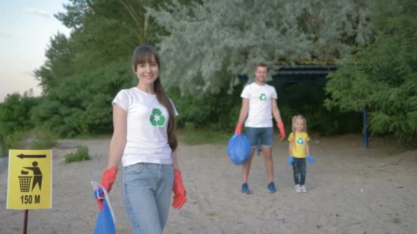 Plajı temizlemek, plastik eldiven takmış, plastik eldiven takmış, arka planda işaret levhası olan genç gönüllü anne portresi. Baba ve küçük kızı plastik çöpleri topladıktan sonra. — Stok video