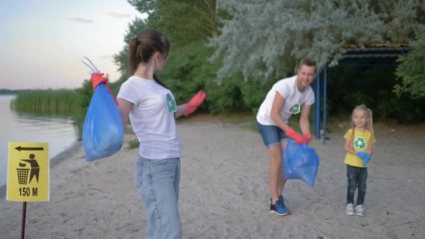 Экологический уход, портрет веселой активистки в резиновых перчатках с мешком для мусора рядом с указателем на заднем плане мужчины с маленькой девочкой, собирающей пластиковый мусор на набережной — стоковое видео