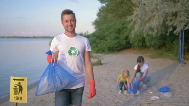 Situación ecológica, retrato de un joven voluntario en guantes de goma con bolsa de basura cerca del cartel de puntero en el fondo hembra con niña recogiendo basura de plástico en la playa — Vídeo de stock