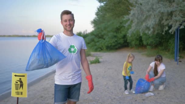 Ecology rescue, portrait of smiling activist man in rubber gloves with garbage bag near pointer sign on background wife with little daughter collecting plastic litter on embankment — ストック動画