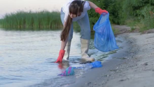 Environmental problems, young activist girl into rubber boots cleans waterfront from plastic rubbish and collects polyethylene trash in garbage bag while standing — ストック動画