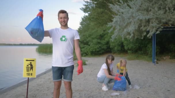 Захист навколишнього середовища, портрет щасливого добровольця чоловічої статі в гумових рукавичках зі сміттєвим мішком біля знака покажчика на фоні жінки з дитиною, що збирає пластикові відмови на пляжі — стокове відео