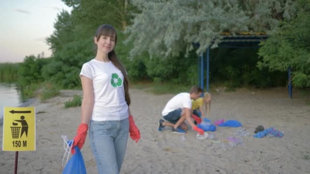 Экологические решения, портрет красивой женщины-активистки в резиновых перчатках с мешком для мусора рядом с указателем на заднем плане мужчина с девочкой, собирающей пластиковые отходы на набережной — стоковое видео