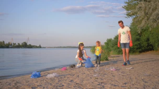 Család önkéntesek, fiatal apa és anya kislányával gyűjti hulladékot szemeteszsákban a piszkos strandon műanyag, miközben takarítás a folyó közelében — Stock videók