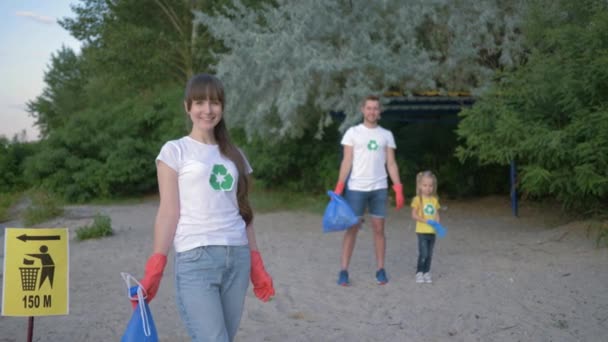 Сохранить планету, портрет молодой женщины-волонтера в резиновых перчатках с мешком для мусора возле указателя знак на заднем плане мужчина и маленькая девочка после сбора пластиковых отбросов на пляже — стоковое видео