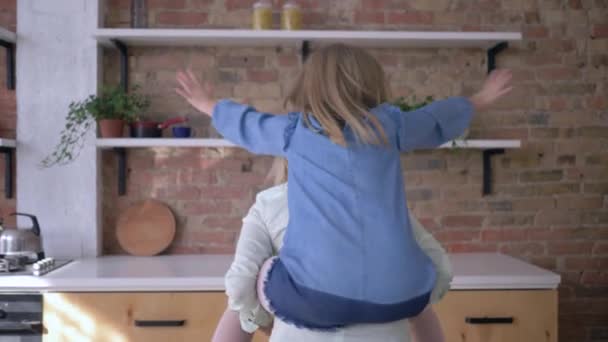Игры с матерью, смешная маленькая девочка играет с мамой сидит на спине и делает самолет руки вытянуты в семейной кухне — стоковое видео