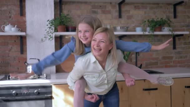 Se divertindo com a mãe, alegre menina brinca com a mãe sentada nas costas e faz plano de braços estendido na cozinha — Vídeo de Stock