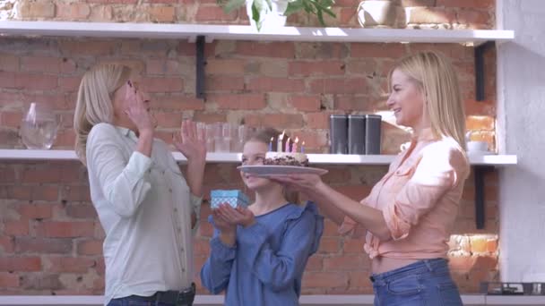 Sorpresa durante Compleanno di Madre, le figlie piccole e adulte con regalo e torta di vacanza con candele si congratulano con la mamma in cucina di famiglia — Video Stock