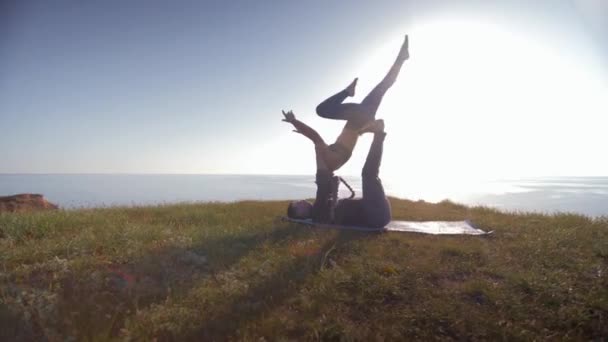 Yoga, acróbata atlética femenina se equilibra en las piernas y las manos de su pareja masculina en la luz del sol sobre el fondo del cielo y el mar en la montaña — Vídeo de stock