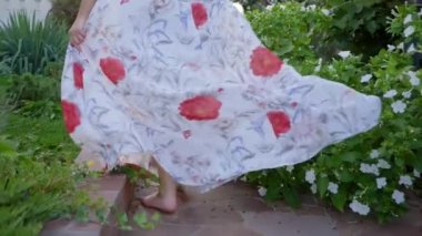 Uzun elbiseli çıplak ayaklı kız çiçeklerin ve yeşilliklerin arasında koşuyor.