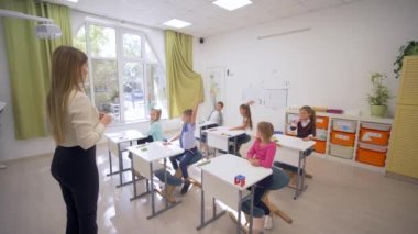 Bir grup öğrenci ders sırasında cevap vermek için el kaldırırken, okulda öğretmenlerin önünde tahtaya oturuyorlar.