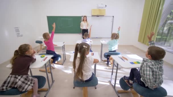 Группа школьников поднимает руки, чтобы ответить на урок, сидя за столом перед учителем на борту в начальной школе — стоковое видео