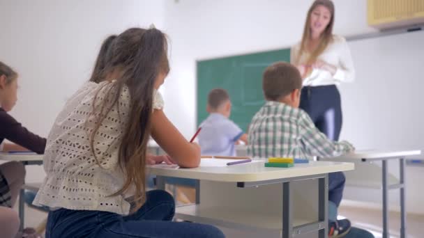 Portret uśmiechniętej uczennicy przy biurku podczas lekcji w klasie w szkole podstawowej na nieskupionym tle — Wideo stockowe