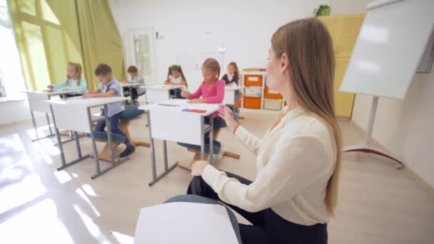 Portret uśmiechniętej nauczycielki podczas lekcji z uczniami w klasie w szkole podstawowej na nieskupionym tle — Wideo stockowe