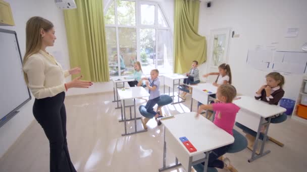 Професійна вчителька розмовляє з маленькими милими дітьми учнями за столами під час уроку освіти в класі в школі — стокове відео