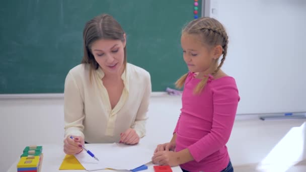 Children schooling, smiling woman educator checks homework at little learner girl at table near blackboard in classroom of school — ストック動画