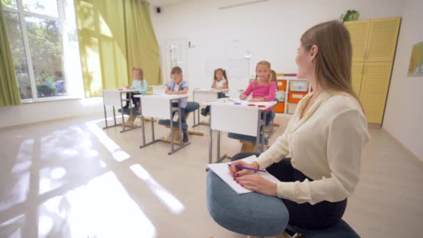 Retrato del feliz profesor sonriente durante la lección sobre los antecedentes de los alumnos niños pequeños detrás de los escritorios en la escuela — Vídeo de stock