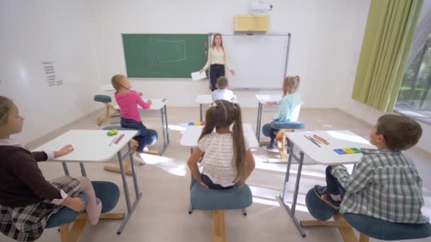 Skupina školáků zvedne ruce, aby odpověděli na lekci, zatímco sedí za stolem před pedagogem na tabuli v juniorské škole — Stock video