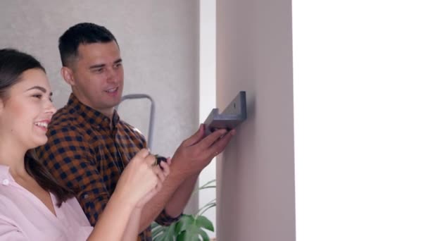 Repair in house, young couple with tape measure in hands hanging shelf on wall during renovation in apartment — ストック動画