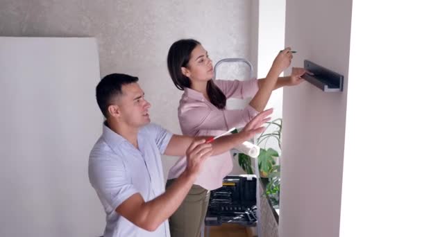 Joven esposa y marido con destornillador cuelgan estante en la pared durante la reparación en el apartamento — Vídeos de Stock