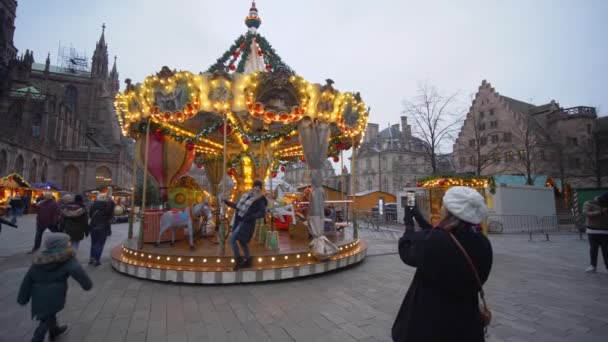 Festive atmosphere in tourist town, children Christmas carousel in garlands and tourists are photographed on mobile phone on city square — Stock Video