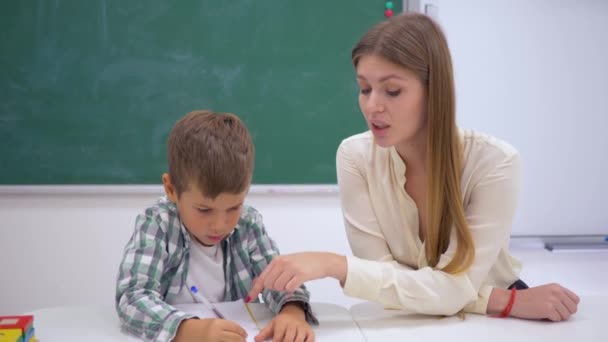 Elementary school, professional tutor helps learning to write to schoolchild at table near board — Stock Video