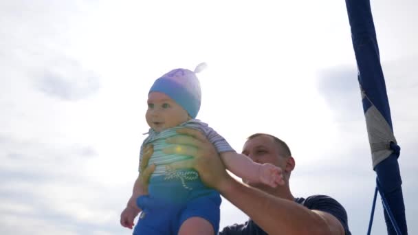 Papá crió al bebé en brazos, niño pequeño en padres manos fuertes en la luz del sol, niño en manos de papá en el fondo azul del cielo , — Vídeos de Stock