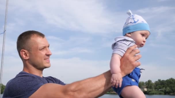 Little boy in fathers strong hands in blue sky background, beautiful male raises son up in arms on lake, — Stock Video