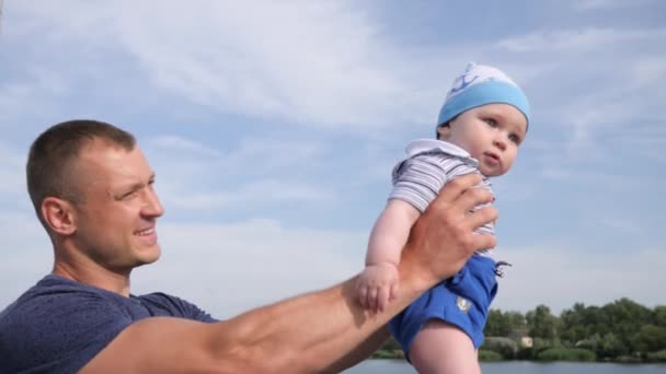 Lucky male raises son up in arms on lake, happy daddy holds into hand first child near river, beautiful dad play with baby — Stock Video