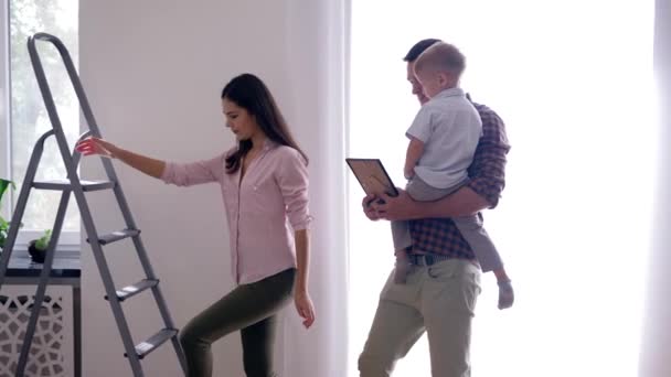 Praca zespołowa remontu mieszkania, szczęśliwa rodzina z dzieckiem wykonująca naprawy w mieszkaniu i powieszenie malarstwa za pomocą drabinki schodowej w domu — Wideo stockowe