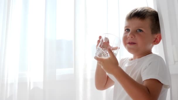 Glas vatten i händerna på liten pojke som dricker mineralvatten på bakgrund vita gardiner — Stockvideo