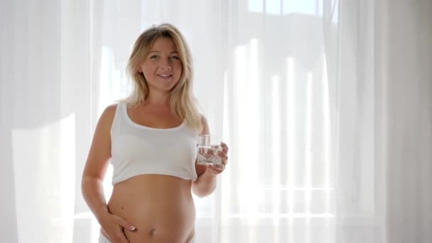 Lady in white T-shirt drinks fresh water in glass and caressing naked belly near window — Stock Video